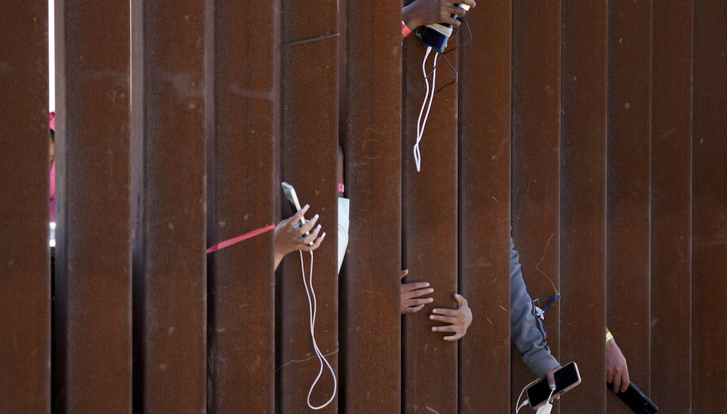 Un grupo de migrantes esperando a solicitar asilo en Estados Unidos extienden sus celulares a través de barreras fronterizas el 11 de mayo de 2023 en San Diego. (AP)