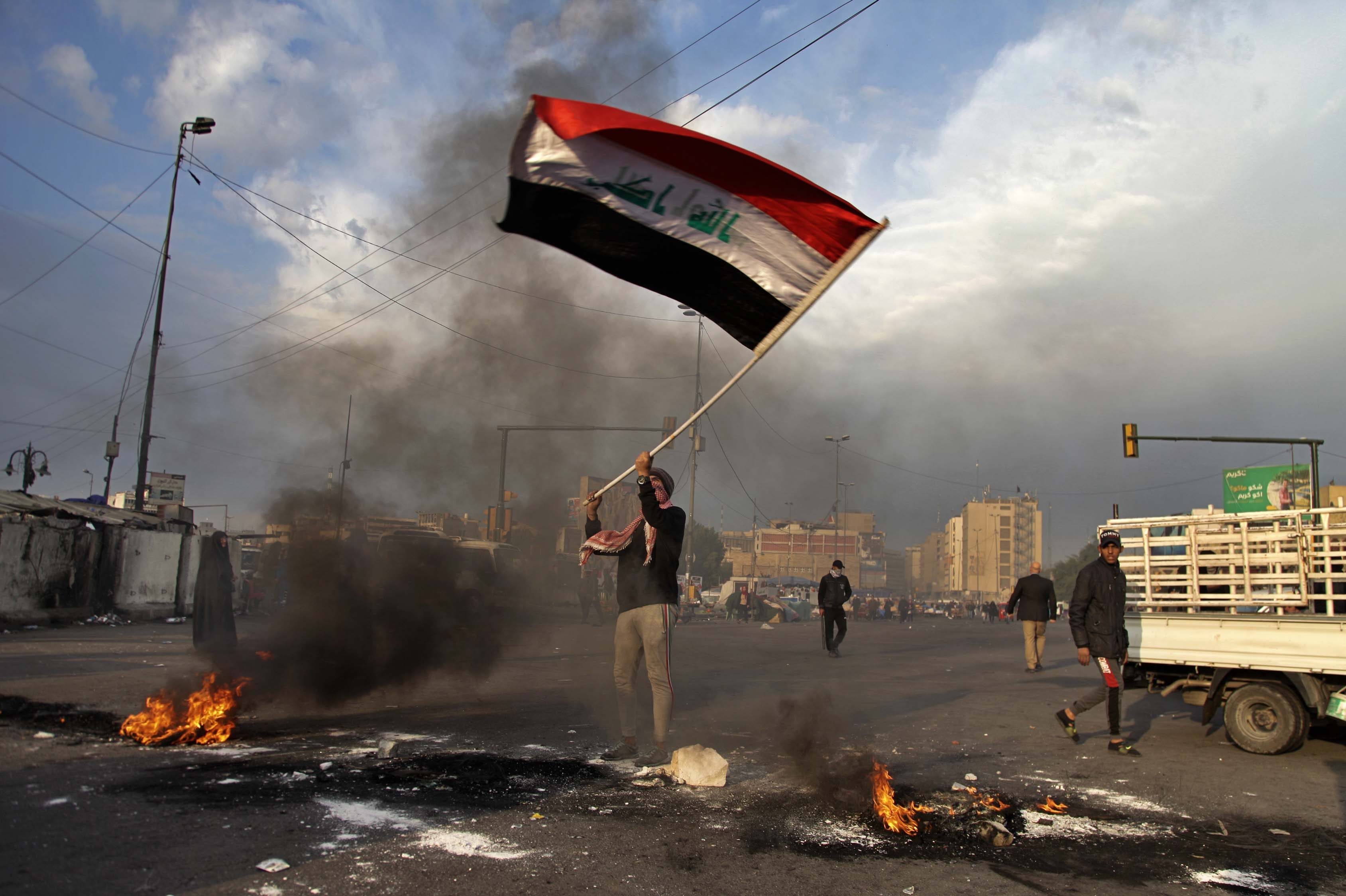 A protester waves the national flag while demonstrators set fire to close streets near Tahrir Square during a demonstration against the Iranian missile strike in Baghdad, Iraq on Jan. 8, 2020. (AP)