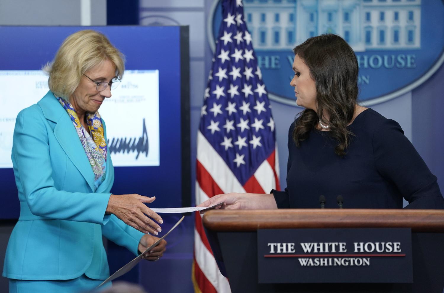 White House press secretary Sarah Huckabee Sanders, right, hands Education Secretary Betsy DeVos, left, a check signed by President Donald Trump in the Brady Press Briefing room of the White House in Washington, Wednesday, July 26, 2017. (AP)
