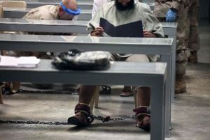 This 2010 photograph, made through one-way glass and reviewed by the U.S. military, shows a shackled Guantanamo detainee reading his materials in a "Life Skills" class as guards behind him shackle another detainee to the floor after bringing him to class.